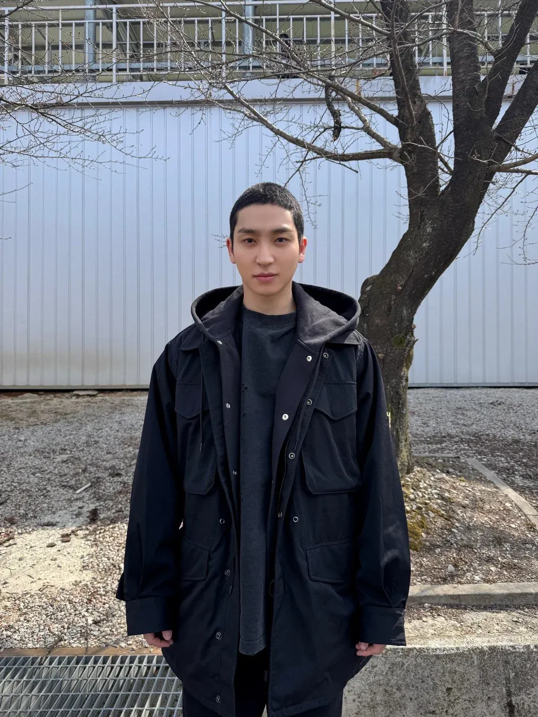 Sangyeon of THE BOYZ with short buzzed hair, wearing an oversized black hooded jacket, standing outdoors near a bare tree.